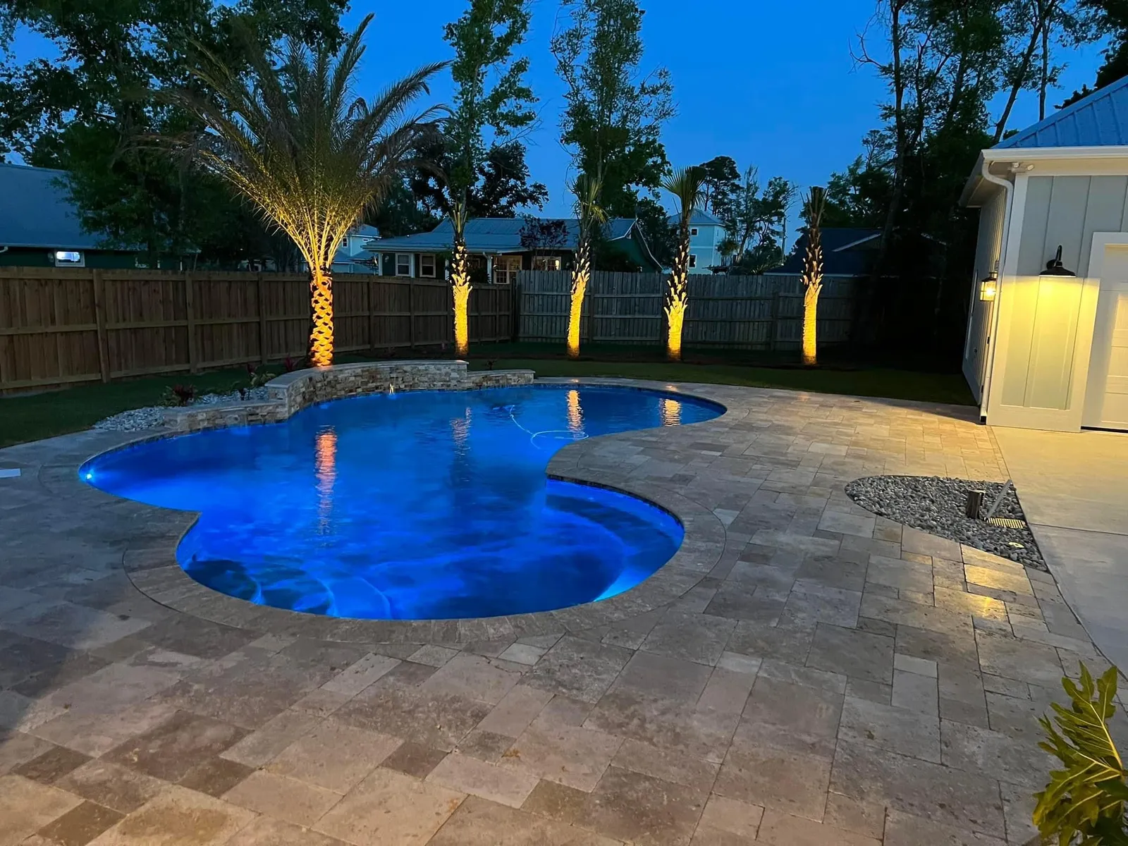 A lit pool at dusk ringed by travertine pavers, with uplit palms along a privacy fence behind it
