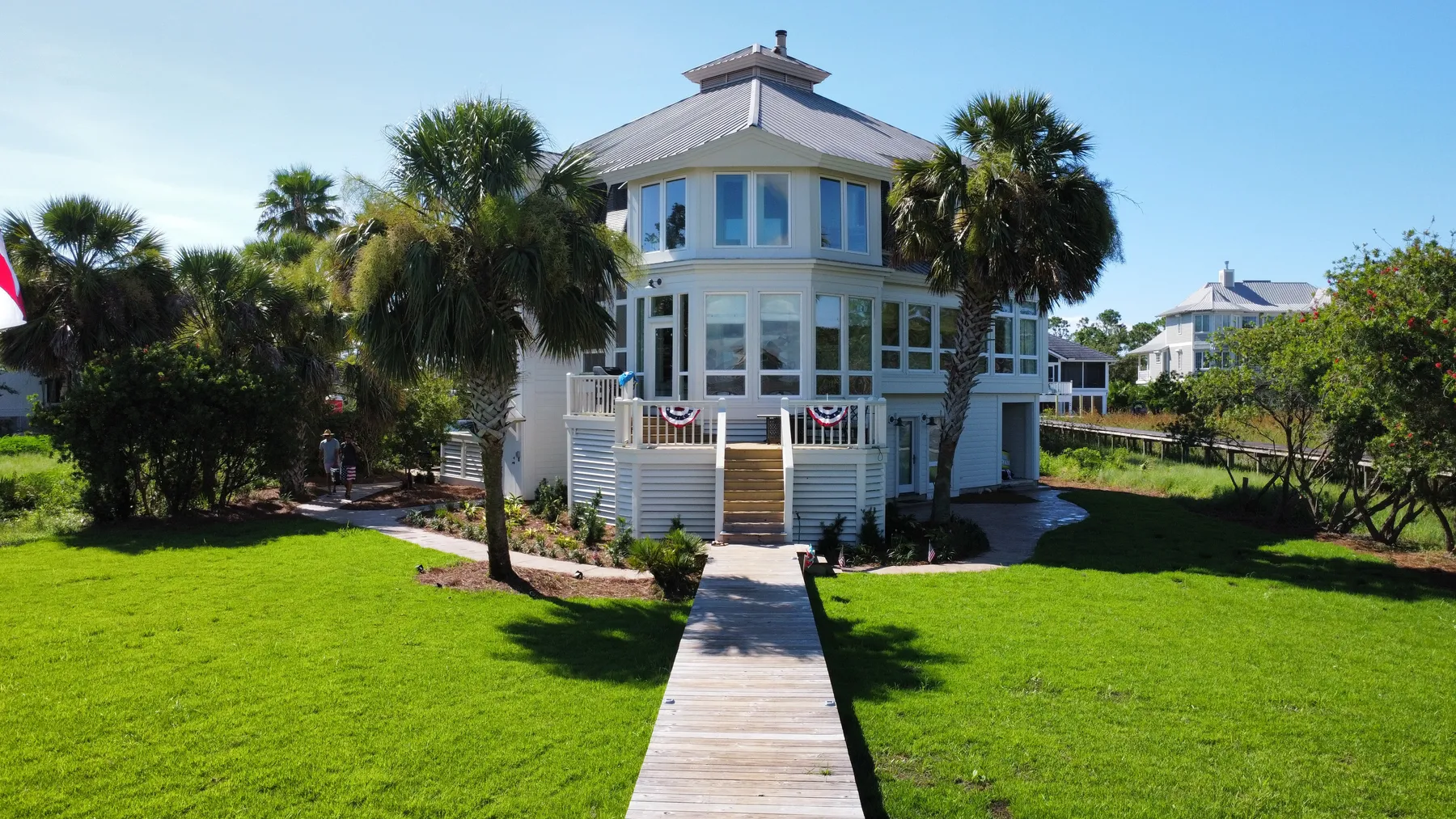 A tall white beach house with a lantern top, a boardwalk path through a mowed lawn and palm clusters on each side